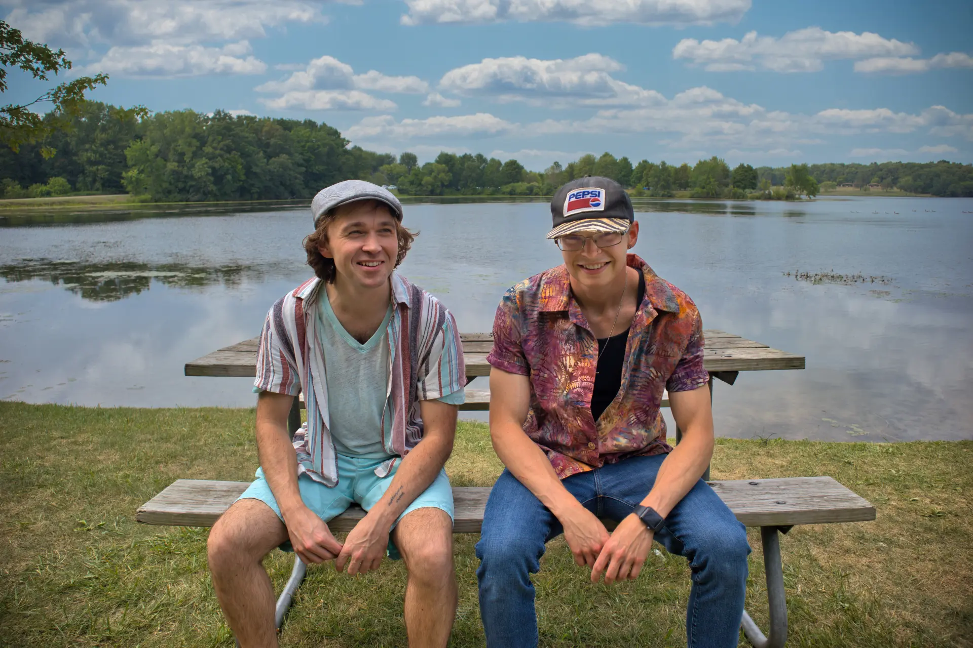 Of the band COAST, Stephen Michael and Jake Long sitting at a picnic table beside a lake on a sunny day, smiling.